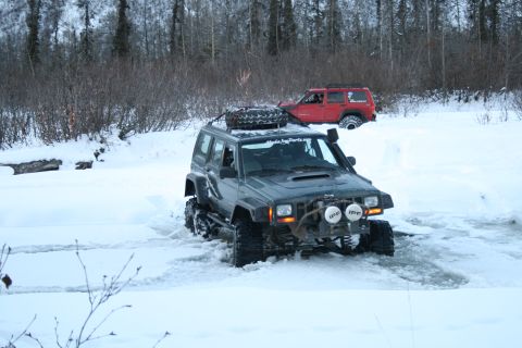 2003 Jeep TJ - A green Jeep Cherokee navigates through a snowy landscape, with another red Jeep visible in the background.