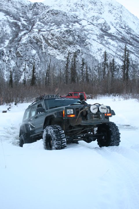 2003 Jeep TJ - A modified dark green SUV navigates through deep snow, with a red vehicle visible in the background. The vehicle appears to be a Jeep Chero…