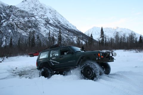 2003 Jeep TJ - A modified Jeep Cherokee XJ navigates a snowy trail with a red pickup truck in the background.