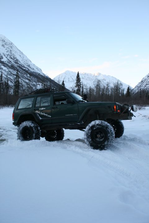 2003 Jeep TJ - A modified dark green Jeep Cherokee XJ navigates a snowy landscape with snow-capped mountains in the background.