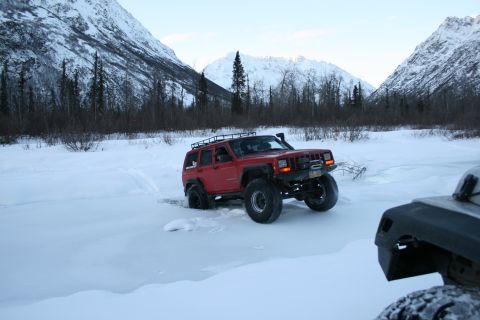2003 Jeep TJ - A red Jeep Cherokee navigates a snowy landscape, partially obscured by another vehicle in the foreground.