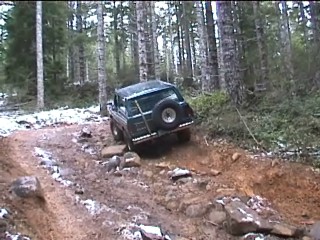 1979 Ford Bronco - A dark green SUV tackles a challenging, rocky trail through a forest, with snow patches visible on the ground.