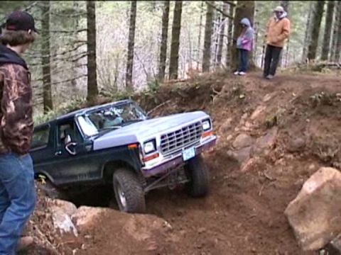 1979 Ford Bronco - A dark blue and silver Ford Bronco tackles a challenging off-road course with spectators observing.