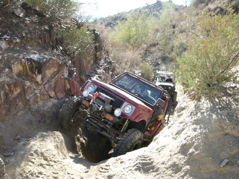 1987 Suzuki Samurai - Two off-road vehicles tackle a challenging, narrow path through a rocky, desert-like landscape.