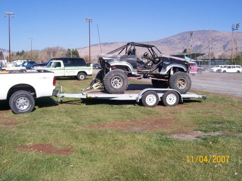 1984 Ford Bronco - Modified SUV on trailer, towed by white pickup truck. Other vehicles visible in background.