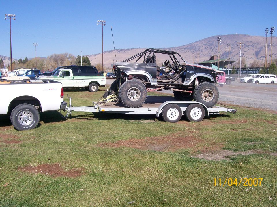 Modified off-road SUV with roll cage on trailer, towed by white pickup. Another white pickup and a white SUV are in the background