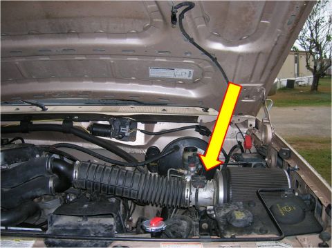 1996 Ford Bronco - Engine bay of a tan/gold Ford Bronco with the hood raised, showing the black ribbed air intake tube running to a cylindrical air filter hou…