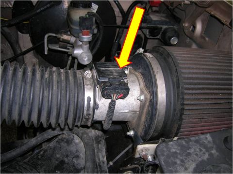 1996 Ford Bronco - Close-up of an engine bay showing an aluminum mass air flow (MAF) sensor housing with a black wiring connector, indicated by a yellow arrow…