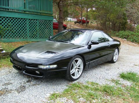1995 Ford Probe - A black 1995 Ford Probe is parked outside on a gravel driveway, with a green structure to its left and trees in the background.