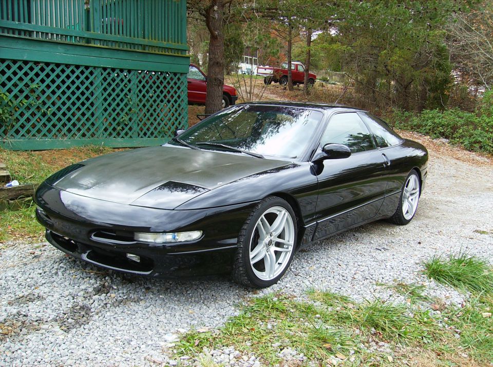 Black 1995 Ford Probe parked on gravel in front of a green lattice-covered structure. Two red trucks are visible in the background