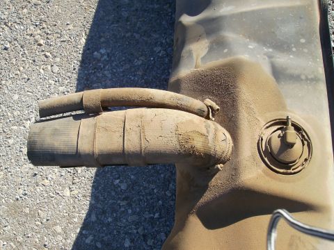 1979 Ford F-100 - A dusty fuel tank with two hoses attached and a fuel cap visible. The part is shown outdoors on a gravel surface.