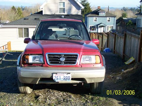 1988 Suzuki Samurai - Front view of a red SUV with a damaged headlight and bumper, parked in a residential area.