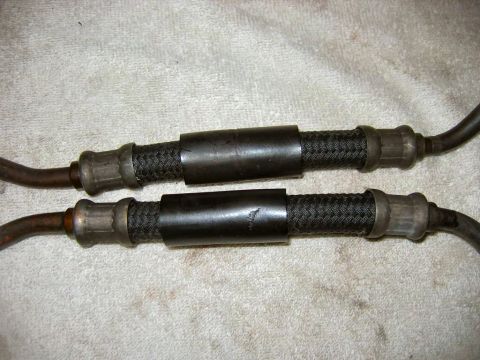 1985 Ford LTD Crown Victoria - Close-up view of two braided brake hoses with metal fittings and protective sleeves, laid out on a textured surface.