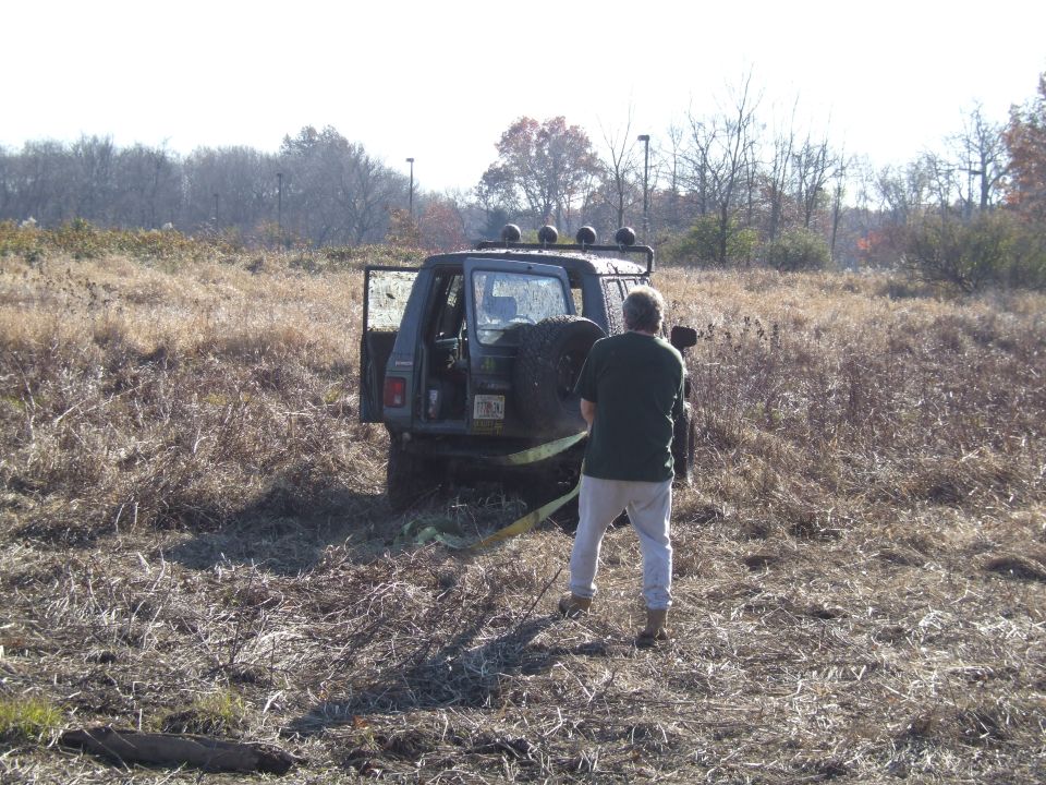 guy trying to pull out a dark SUV stuck in tall dry grass, spare tire mounted on back