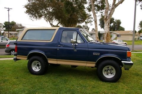 1986 Ford Bronco - A blue Ford Bronco with tan trim is parked on a grassy lawn in front of houses.