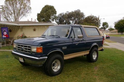 1986 Ford Bronco - A dark blue and tan Ford Bronco is parked on a lawn in front of a house.
