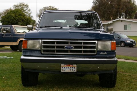 1986 Ford Bronco - A dark blue Ford Bronco is parked on a grassy lawn in front of a house. Another vehicle is visible to the left.