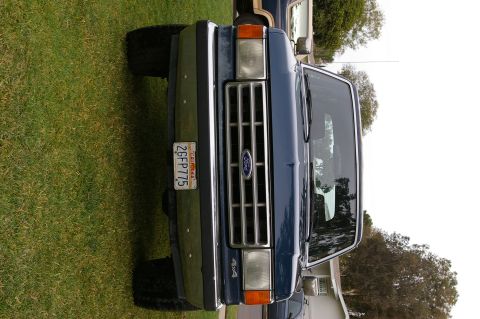 1986 Ford Bronco - Front view of a blue Ford Bronco with a 4x4 badge, parked on grass next to another vehicle.
