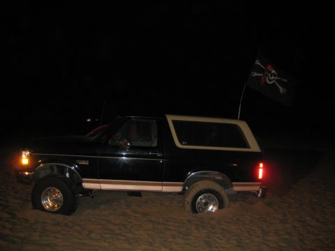 1994 Ford Bronco - Black Ford Bronco with tan top stuck in sand at night, flying a pirate flag.