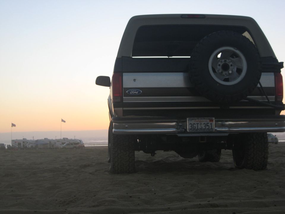 1994 Ford Bronco rear view with spare tire mounted on the back, parked on sand with RVs and flags in the distance