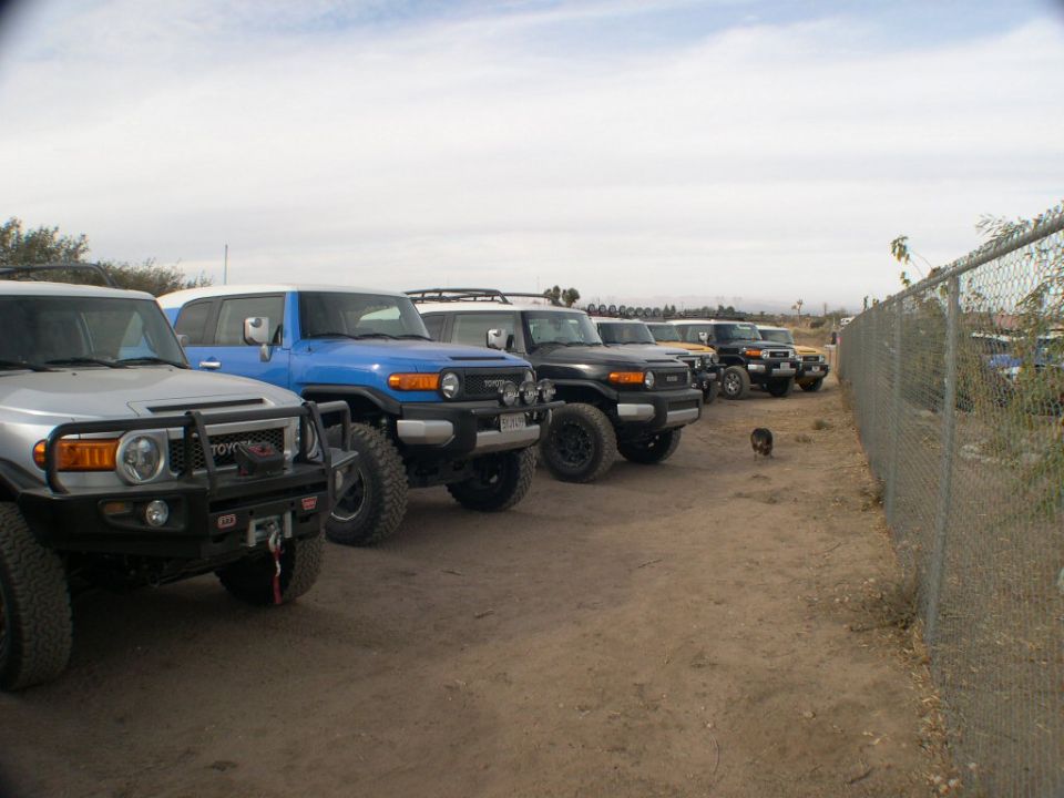 Several Toyota FJ Cruisers parked in a dirt lot next to a fence. One is blue, one is silver, and others are black and yellow