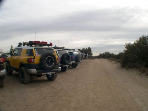 2007 Toyota FJ Cruiser - A line of Toyota FJ Cruisers, including a yellow one with a roof rack, are parked on a dusty trail under an overcast sky.