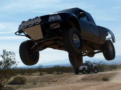 2007 Ford Ranger - A modified truck soars through the air during an off-road event, with another vehicle visible on the dusty track below.