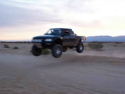 2007 Ford Ranger - A black off-road pickup truck soars through the air during a desert jump, with mountains visible in the background.