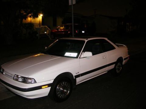 1991 Mazda MX-6 - A white 1991 Mazda MX-6 coupe is parked on a street at night, with a red car visible in the background.