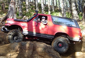 1986 Ford Bronco - This red Ford Bronco is equipped for off-road adventures, navigating a rocky trail in a wooded area.