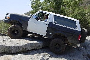 1986 Ford Bronco - A black and white Ford Bronco navigates a rocky terrain on a sunny day.
