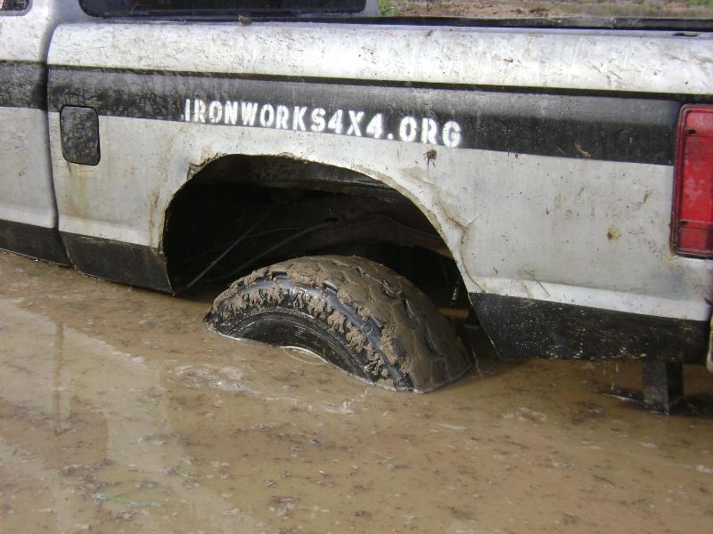 Rear wheel of a silver pickup buried in mud below an IRONWORKS4X4.ORG bedside decal