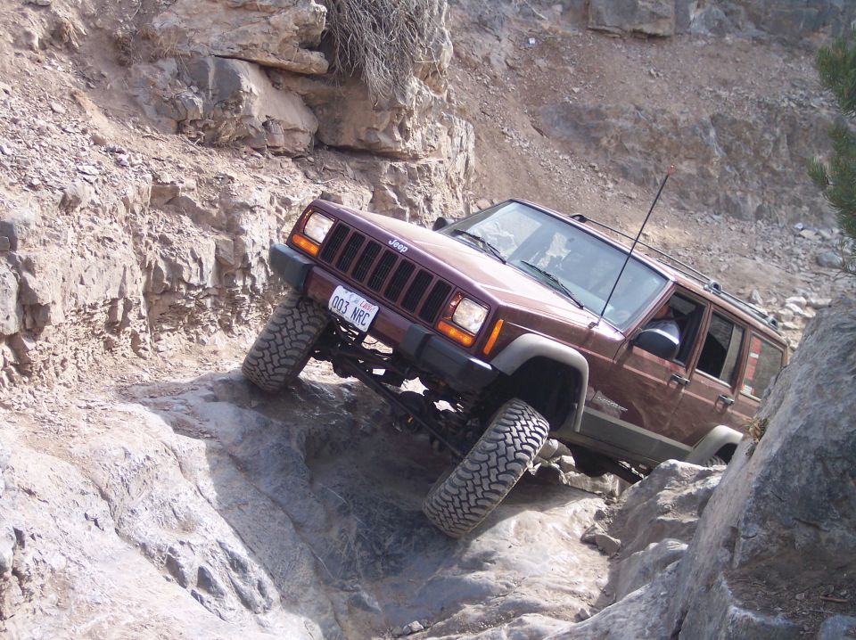 Maroon Jeep Cherokee XJ with lifted suspension and off-road tires navigating a rocky trail. License plate reads 003 NRC
