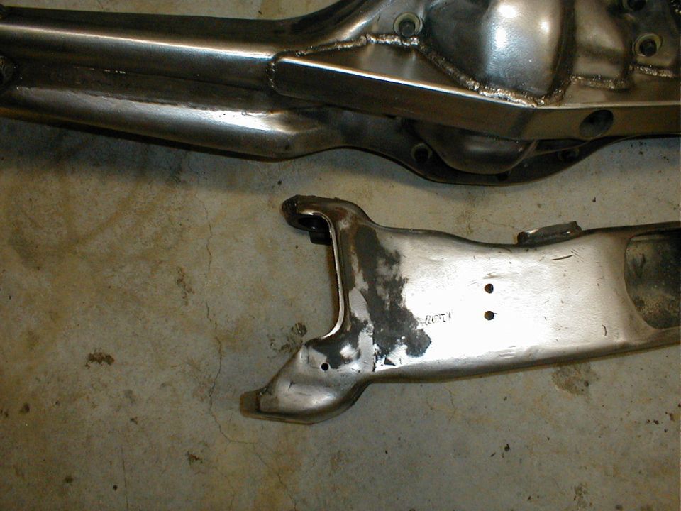 Two metallic suspension parts on a concrete floor. One is a long, tubular component with welded seams. The other is a curved, cast metal piece with two holes