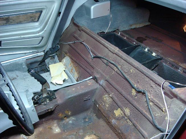 Stripped car interior showing rusty bare floor pan, seat belt, wiring, and removed carpet during restoration