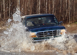 1986 Ford Bronco - A dark blue Ford SUV blasts through a deep water crossing on a dirt trail, throwing a large wall of spray off the driver's side. Bare leafl…