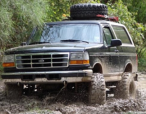 1986 Ford Bronco - This black Ford Bronco is shown from a three-quarter front angle, navigating a muddy off-road trail.