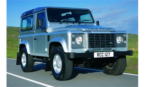 1989 Ford Bronco - A silver Land Rover Defender 90 is captured in a three-quarter front view while driving on a paved road under a clear blue sky.