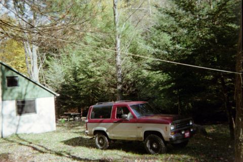 Two-tone red and tan Ford Bronco II parked on grassy area next to a cabin