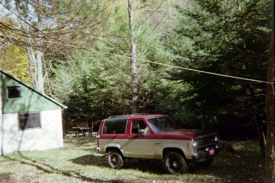 Two-tone red and tan Ford Bronco II parked on grassy area next to a cabin