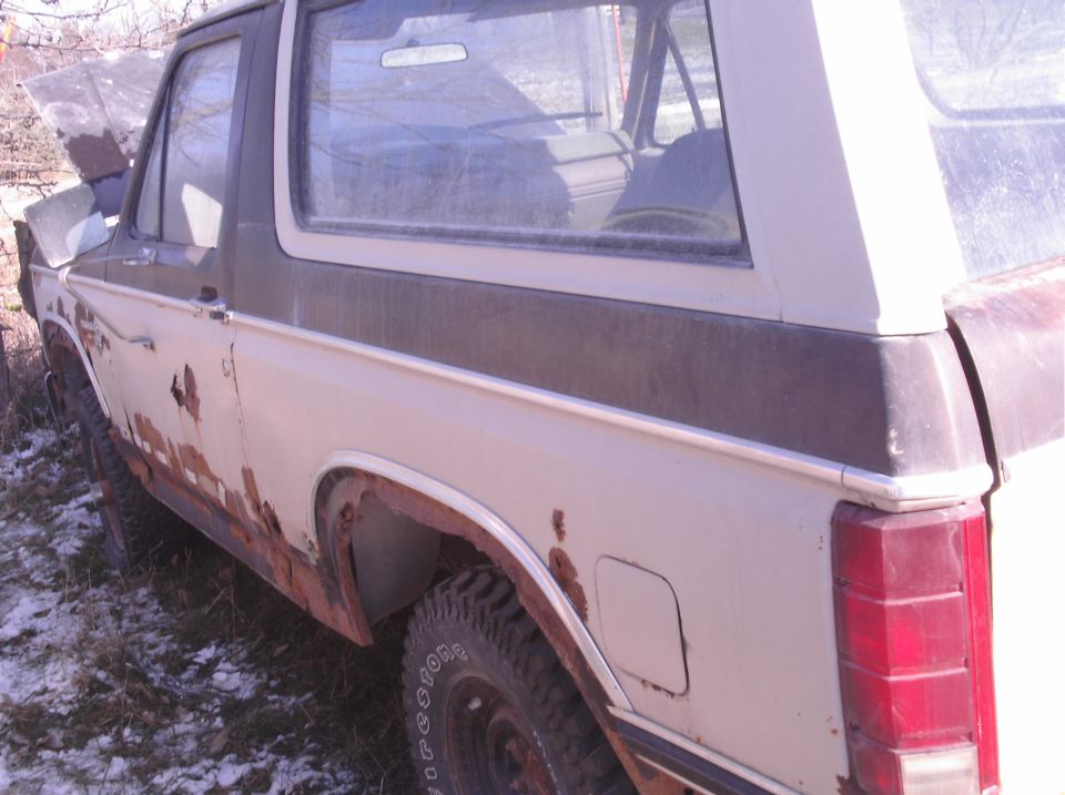 Side view of a 1980 Ford Bronco with significant rust on the lower body panels and hood open
