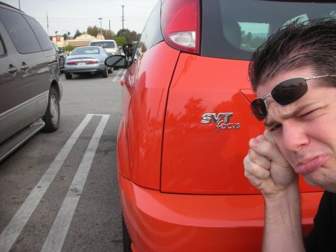 1998 Ford Crown Victoria - man makes a pained face leaning against the rear of a red Ford SVT Focus showing its SVT badge in a parking lot.