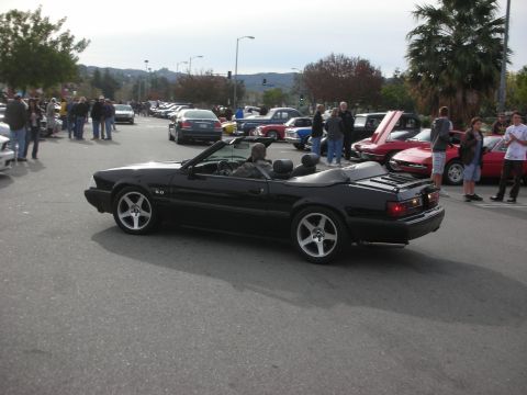 1998 Ford Crown Victoria - black Fox-body Ford Mustang 5.0 convertible drives slowly through a crowded outdoor car show.