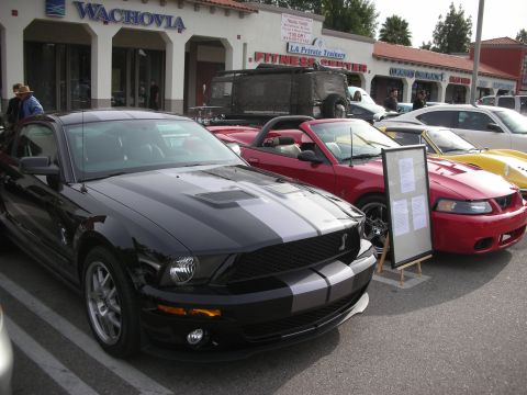 1998 Ford Crown Victoria - black Mustang with silver racing stripes parked next to a red convertible in front of storefronts, with an event sign on an easel.