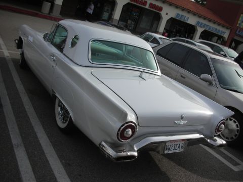 1998 Ford Crown Victoria - Rear three-quarter view of a white classic Ford Thunderbird hardtop with porthole window and round taillights, parked outside shops.