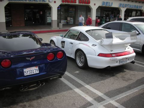 1998 Ford Crown Victoria - white Porsche 911 track car with number 27 and a tall rear wing parked next to a blue Corvette coupe in front of storefronts.