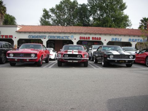 1998 Ford Crown Victoria - Front view of three classic muscle cars, two Camaros flanking a red-striped Mustang, parked in front of storefronts at a car show.