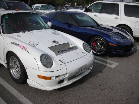 1998 Ford Crown Victoria - white widebody Porsche 911 with Octane Gallery livery parked beside a blue Chevrolet Corvette in a lot.