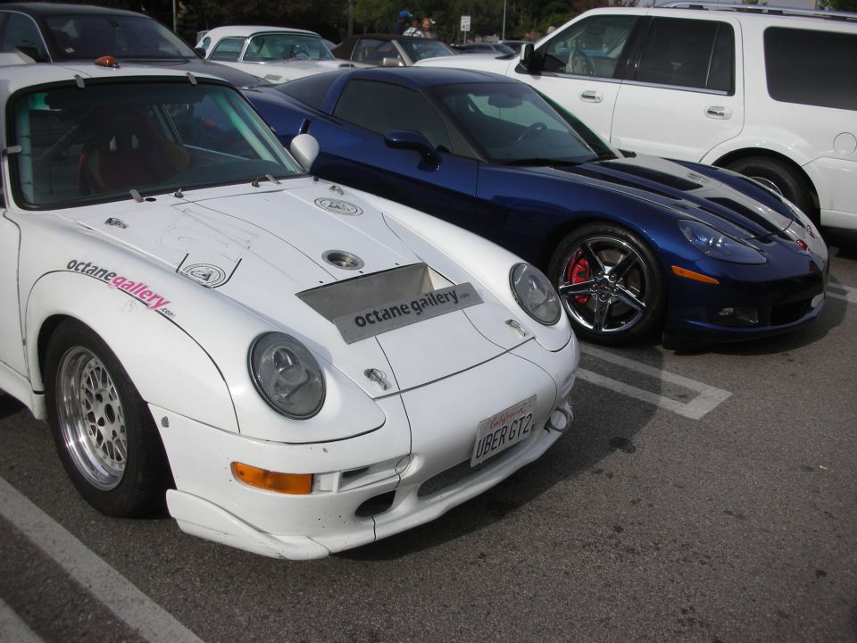 White widebody Porsche 911 beside a blue Chevrolet Corvette at a car meet