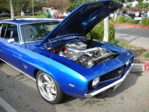 1998 Ford Crown Victoria - blue first-generation Chevrolet Camaro with hood up, revealing a polished supercharged engine, at an outdoor show.